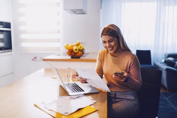 virtual - care for employers - woman in sweater holding bills in one hand and in other smart phone. On table are laptop and bills